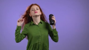 Müzik dinleyen Avrupalı bir bayan, kablosuz taşınabilir hoparlörle dans ediyor - modern ses sistemi. Uzun kızıl saçlı bir kadın Violet stüdyosunun arka planında eğleniyor. Müziğin ritmine göre hareket eder..