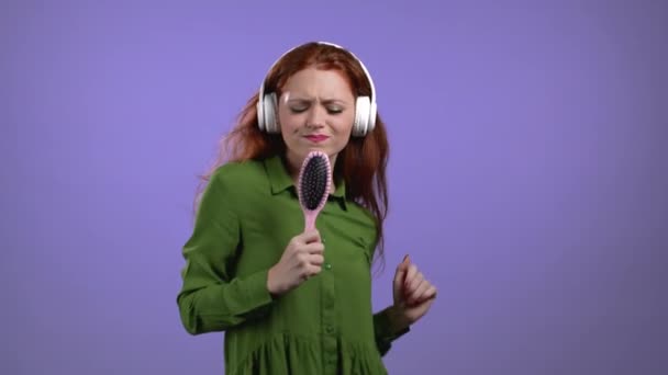 Европейская женщина поет и танцует с расчёской или расчёской вместо микрофона на фоне фиолетовой студии. Леди в наушниках развлекается, слушает музыку, мечтает стать знаменитостью. — стоковое видео