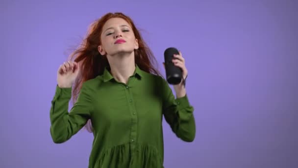 Европейская леди слушает музыку, танцует с беспроводным портативным динамиком - современная звуковая система. Женщина с длинными рыжими волосами наслаждается на фоне фиолетовой студии. Она движется к ритму музыки. — стоковое видео