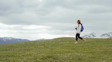 Genç gezgin kadın yüksek dağ vadisinde tek başına yürüyor. Sırt çantası ve kamerası olan kadın. Komşu kayaların karla kaplı zirveleri. Yavaş çekim.