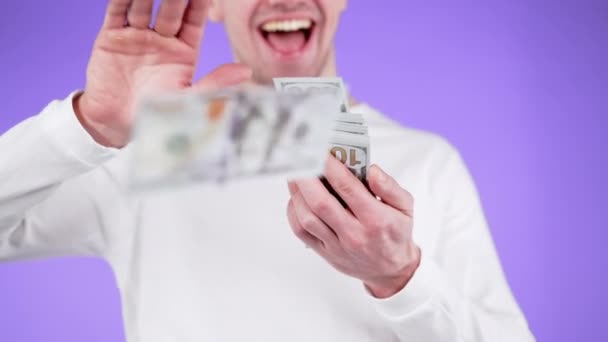 Jongeman met een glimlach verstrooit geld. Onherkenbare man besteedt te veel geld aan USD. Guy is flush met dollars op violet studio achtergrond. — Stockvideo