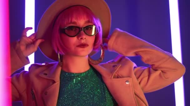 Эксцентричная женщина с розовыми волосами, позирующая рядом с цветными неоновыми лампами. Очаровательная странная девушка в шляпе и солнцезащитных очках, концепция ночной жизни. Современный модельный поп-наряд, стиль жизни. — стоковое видео