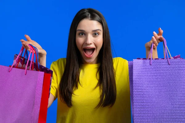 Aufgeregte Frau mit bunten Papiertüten nach dem Einkauf auf blauem Studiohintergrund. Konzept des saisonalen Verkaufs, Käufe, Ausgaben für Geschenke — Stockfoto