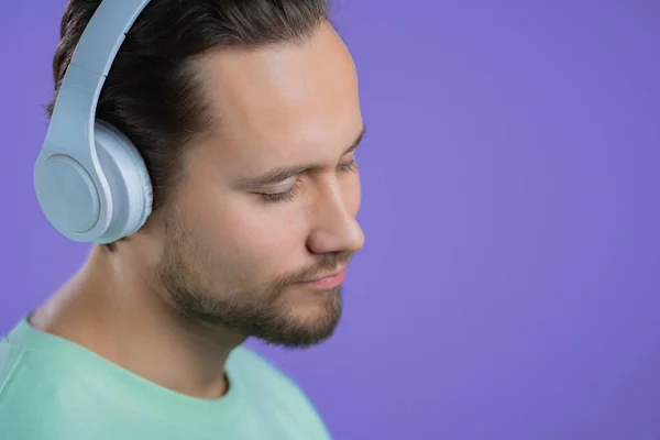 Jeune homme écoutant de la musique avec des écouteurs sans fil, gars souriant en studio sur fond violet. concept radio. — Photo