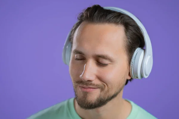 Ung man lyssnar på musik med trådlösa hörlurar, kille ler i studio på lila bakgrund. radiokoncept. — Stockfoto