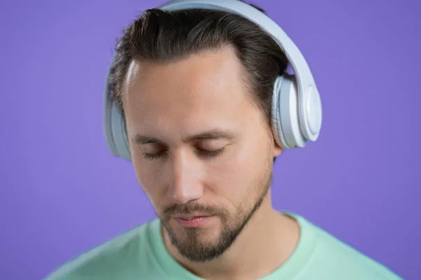 Jeune homme écoutant de la musique avec des écouteurs sans fil, gars souriant en studio sur fond violet. concept radio. — Photo