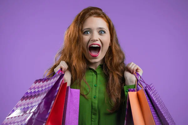Aufgeregte Frau mit bunten Papiertüten nach dem Einkauf auf violettem Studiohintergrund. Konzept des saisonalen Verkaufs, Käufe, Ausgaben für Geschenke — Stockfoto