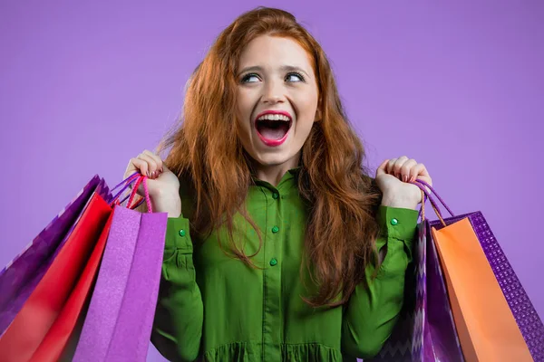 Aufgeregte Frau mit bunten Papiertüten nach dem Einkauf auf violettem Studiohintergrund. Konzept des saisonalen Verkaufs, Käufe, Ausgaben für Geschenke — Stockfoto