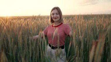 Genç, güzel bir kadın buğday tarlasında kameraya gülümsüyor. Günbatımı arka planı. Kırmızı rujlu ve kısa saçlı kadın. Tarım, doğa, ekoloji kavramı