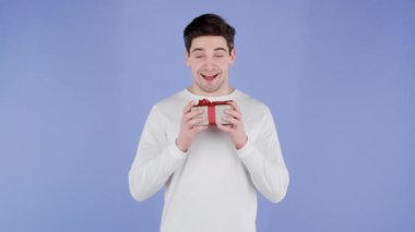 Mor stüdyo arka planında hediye kutusu tutan yakışıklı adam kameraya gülümsüyor. Mutlu Avrupalı adam hediye aldı ve içinde ne olduğuyla ilgilendi..