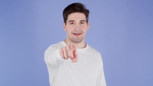 Hombre encantador señala el dedo índice en la lente de la cámara y guiña, coquetea. ¡Eh, tú! Tipo en ropa blanca sobre fondo púrpura. — Vídeos de Stock