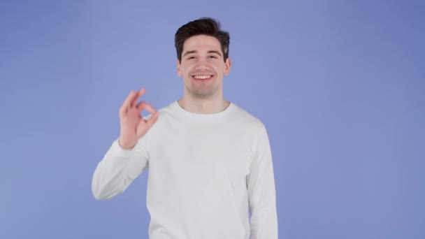 Hombre guapo en ropa blanca muestra bien, ok gesto. Un tipo feliz con fondo violeta. Ganador. Éxito. Modelo masculino positivo sonríe a la cámara. Lenguaje corporal. — Vídeos de Stock