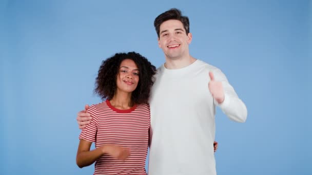 Jovem casal interracial fazendo polegares para cima sinal no fundo azul. Vencedor. Sucesso. Mulher afro-americana positiva e homem branco sorri para a câmera. Linguagem corporal. — Vídeo de Stock