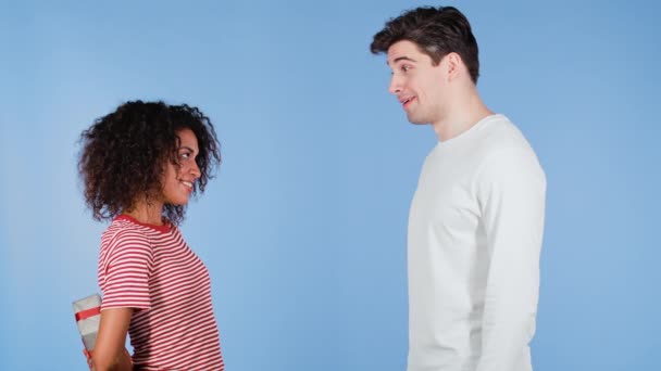 Jong interraciaal koppel op blauwe studio achtergrond. Afro-Amerikaanse vrouw geeft geschenk met rode strik aan haar blanke man of vriendje. Verjaardag, liefde, verrassing, geluk concept. — Stockvideo