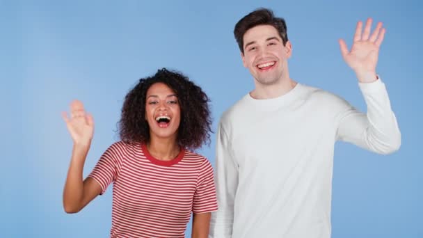 Pareja amigable interracial - hombre blanco y mujer africana saludando con las manos - hola. Saludo, decir hola a la cámara en el fondo del estudio azul. — Vídeos de Stock