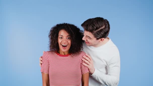 Homem branco fecha os olhos de sua amada namorada afro-americana antes de surpreendê-la. casal interracial em fundo estúdio azul. Amor, férias, conceito de felicidade. — Vídeo de Stock