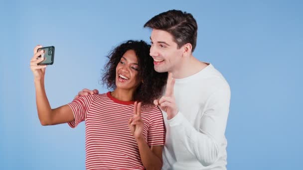 Jovem casal interracial fazendo selfie com smartphone no fundo do estúdio azul. Mulher afro-americana e homem branco sorrindo, se divertindo. Amor, férias, conceito de felicidade. — Vídeo de Stock