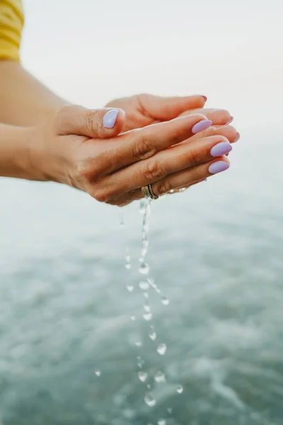 Acqua che sgorga dalle mani delle donne. Mare, vicino alle braccia femminili. Camicia gialla sullo sfondo. L'acqua è una forza vitale — Foto Stock