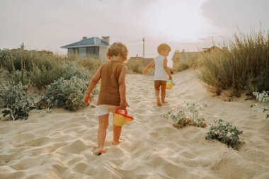 Şirin küçük çocuklar kumlu sahilde bitkileri teneke kutularla suluyor. Yaz güneşli bir gün. Renkli tencereleri olan bebek ikizler. Çocukların doğal estetik portresi.