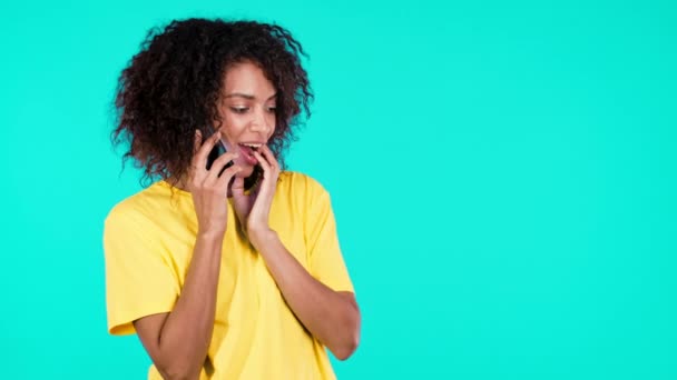 Mujer africana joven habla con sonrisa en el teléfono. Se ríe, noticias sorprendentes o chismes. Chica sosteniendo y utilizando el teléfono inteligente. Teal fondo del estudio. — Vídeos de Stock