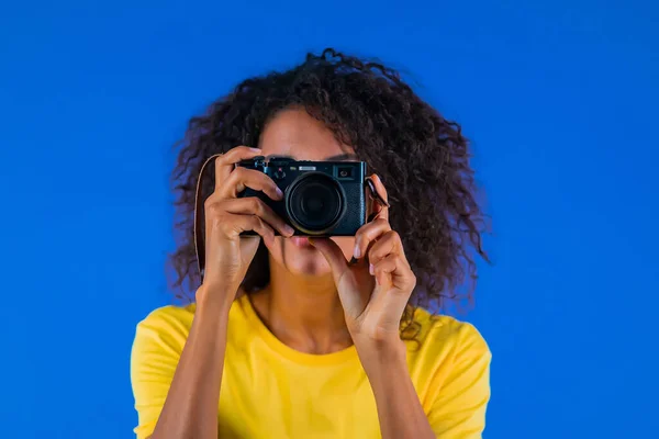 Jovem afro-americana tira fotos com câmera DSLR em fundo azul em estúdio. Menina sorrindo como fotógrafa. — Fotografia de Stock