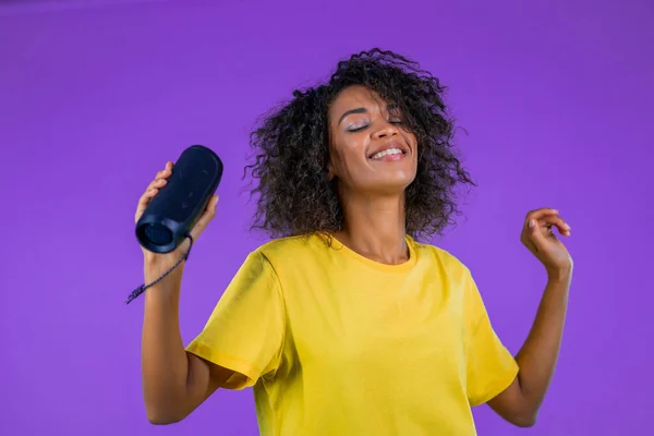 Gemischte Frau hört Musik über drahtlose tragbare Lautsprecher - modernes Soundsystem. Afrikanische Dame tanzt, genießt auf lila Studiohintergrund. Sie bewegt sich im Rhythmus der Musik. — Stockfoto