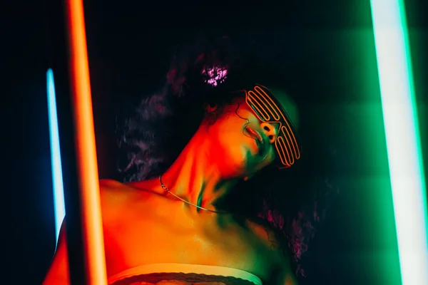 Módní žena s afro účesem tančí v pokoji s barevnými lampami. Cyberpunkový styl nebo hipster s fluorescenčními náramky a brýlemi. Noční klub, futuristický outfit. Sexy dáma koncept. — Stock fotografie