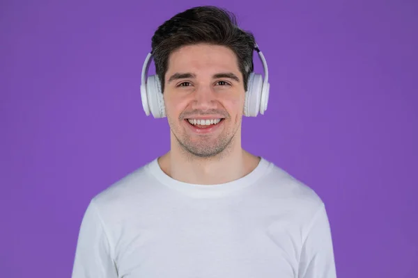 Charmante jongeman die naar muziek luistert met een draadloze hoofdtelefoon, die lol heeft, glimlacht in de studio op een paarse achtergrond. Dans, radio concept. — Stockfoto