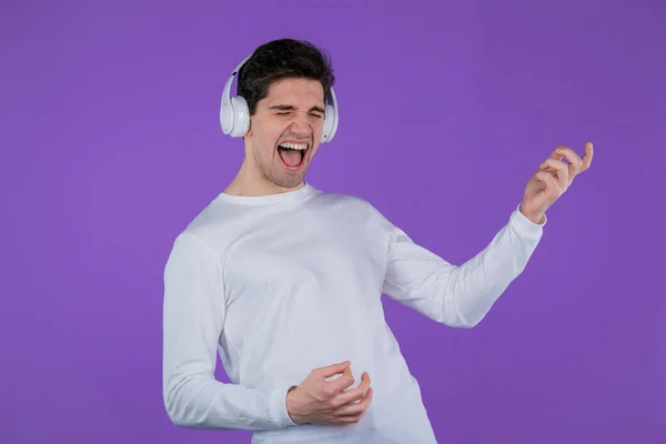 Funny man listening to music with wireless headphones, guy having fun, depicts air guitar playing on purple background. Dance, radio concept. — Stock Photo, Image