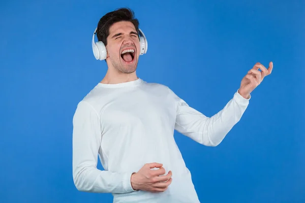 Homem engraçado ouvindo música com fones de ouvido sem fio, cara se divertindo, retrata guitarra de ar tocando em fundo azul. Dança, conceito de rádio. — Fotografia de Stock