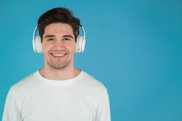 Porträt eines jungen Mannes, der Musik mit drahtlosen Kopfhörern hört, Typ, der im Studio auf blauem Hintergrund lächelt. Tanz, Radio, Kopierraumkonzept. — Stockfoto