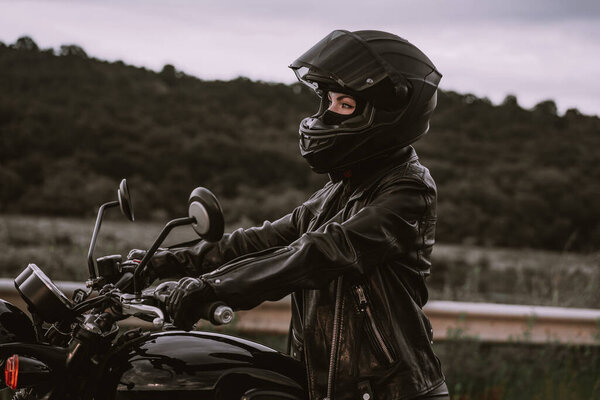 Portrait of confident motorcyclist woman in motorcycle helmet. Young driver biker looking away outdoors alone on highway. Ready for trip. Cafe racers, motorbike aesthetics and vintage design concept.