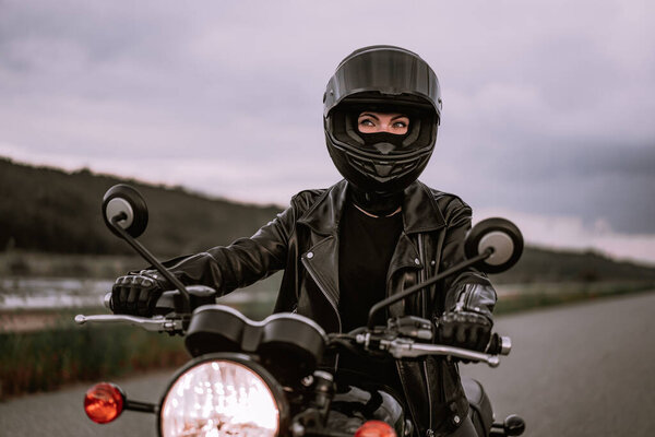 Portrait of confident motorcyclist woman in motorcycle helmet. Young driver biker looking away outdoors alone on highway. Ready for trip. Cafe racers, motorbike aesthetics and vintage design concept.