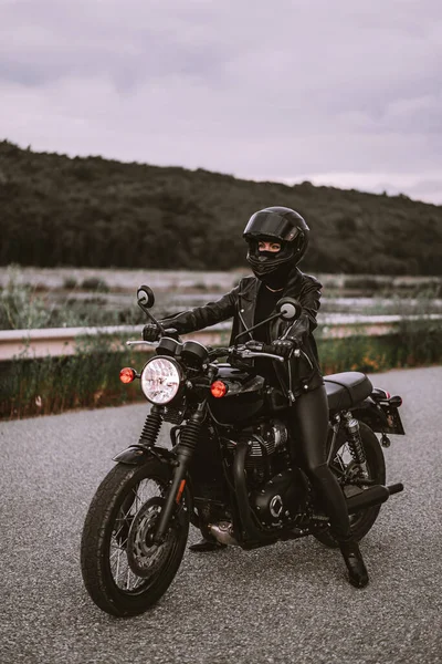 Stilvolle Motorradfahrerin in Helm und Lederjacke sitzt auf einem Oldtimer-Motorrad. Autofahrerin im Freien vor dem Hintergrund der Natur. Reise, Café Racer, Geschwindigkeit, Freiheitskonzept. — Stockfoto