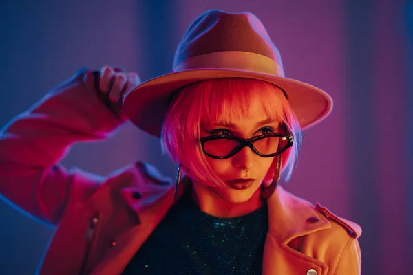 Eccentric woman with pink hair posing under neon light. Charming unusual girl in hat and sunglasses, nightlife concept. Modern model pop outfit, influencer lifestyle. — Stock Photo, Image
