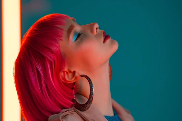 Eccentric woman with pink hair posing near led neon lamp on teal background. Charming odd girl, nightlife concept. Modern model pop outfit, influencer lifestyle. — Stock Photo, Image