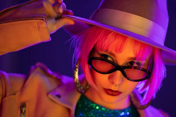 Eccentric woman with pink hair posing under neon light. Charming unusual girl in hat and sunglasses, nightlife concept. Modern model pop outfit, influencer lifestyle. — Stock Photo, Image