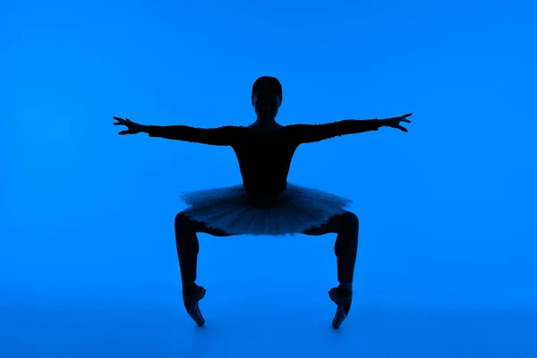 Αγνώριστη μπαλαρίνα με φουστάνι και pointe παπούτσια κάνοντας squat όρθια στις μύτες των ποδιών. Απομονωμένη γυναίκα σε μπλε φόντο. Χορεύτρια μπαλέτου δείχνει κλασικά στοιχεία. — Φωτογραφία Αρχείου