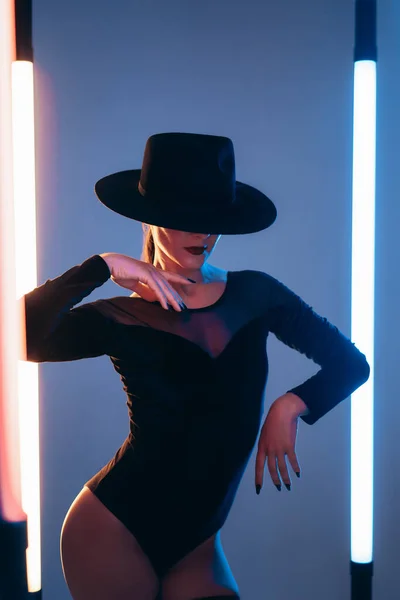 Snygg dansare kvinna i hatt, bodysuit dans med händerna flexibla element. Lady flytta till rytmen av musik mellan neon led glödande lampor på blå bakgrund. — Stockfoto