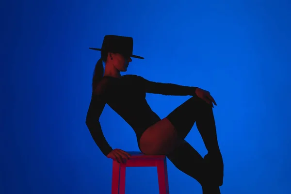 紫色のネオンの下で多色の背景に高い椅子に座って帽子のプロのバレエダンサー。官能的なセクシーなバレリーナはストッキングで彼女の足で踊る。女性のファタールの衣装. — ストック写真