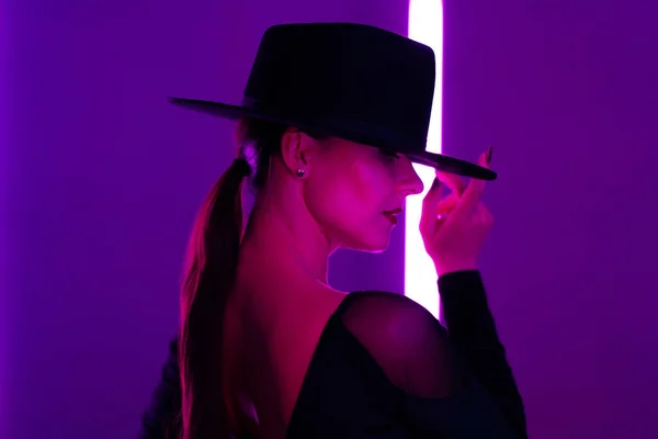 Lockande kvinna i hatt mellan blinkande färgade neonljus, studio bakgrund. Femme fatale, stilren balettdansare. — Stockfoto