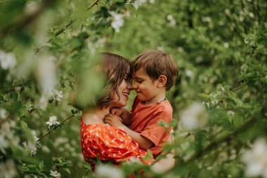 Mutlu anne ve bebeğinin portresi. Anne ve oğlunun güzel dokunaklı sahnesi birbirine hayran ve samimi bir şekilde gülümsüyor. Baharda çiçek açan bahçe. Aile, öpücük, çocukluk kavramı.