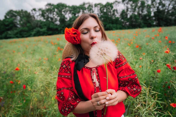 Mulher feliz em blusa vermelha soprando no dente de leão no campo de papoilas. Menina com flor na camisa tradicional ucraniana bordada. Desejando, conceito de alegria — Fotografia de Stock