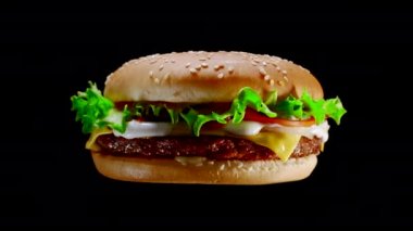 Rotating flying appetizing burger with meat cutlet, onion, vegetables, melted cheese, lettuce and mayonnaise sauce. Isolated hamburger spinning around on black background, close-up view