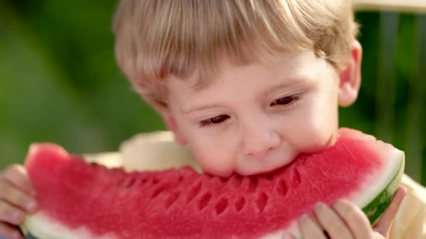 Υπέροχο αγοράκι που τρώει φρέσκο κόκκινο καρπούζι. Νόστιμο βίντεο με ξανθό παιδί κάθεται δίπλα στο τραπέζι στην πίσω αυλή. Υγιεινός τρόπος ζωής, βιταμίνες, καλοκαιρινή συγκομιδή. — Αρχείο Βίντεο