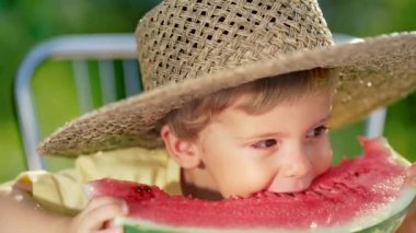 Yakışıklı çocuk, taze kırmızı karpuz yiyor. Gülümseyen bir çocuğun bahçedeki masanın yanında oturduğu enfes görüntüler. Sağlıklı yaşam tarzı, kavun vitaminleri, yaz hasadı..