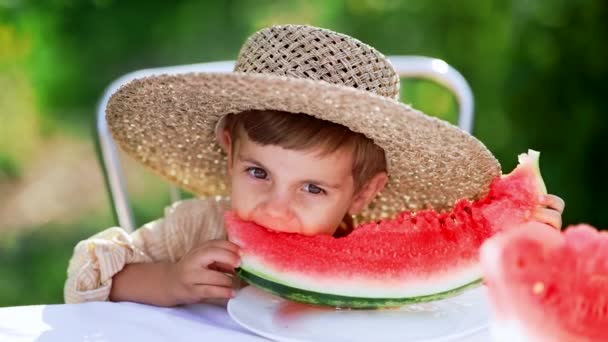 Υπέροχο αγοράκι που τρώει φρέσκο κόκκινο καρπούζι. Yummy πλάνα του χαρούμενου παιδιού σε ψάθινο καπέλο κάθεται στο τραπέζι στην πίσω αυλή. Υγιεινός τρόπος ζωής, βιταμίνες, καλοκαιρινή συγκομιδή. — Αρχείο Βίντεο