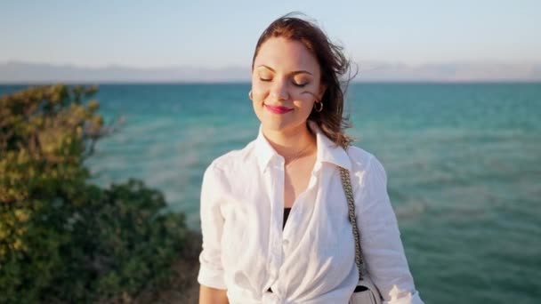Mosolygó nő portréja, a sziklafalon áll, élvezi a szelet, a friss levegőt. Kék tengeri háttér. Fehér inges hölgy. Utazási cél, nyaralás Görögországban. — Stock videók