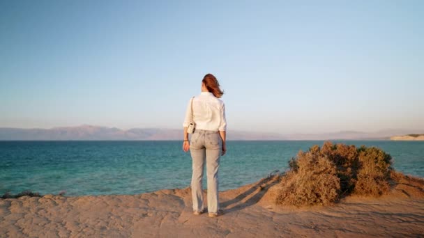 Mujer irreconocible parada cerca del acantilado sobre el mar Jónico Mediterráneo. Señora observando hermosa superficie de agua azul solo, fondo de la naturaleza. Clima ventoso, hora dorada. — Vídeo de stock