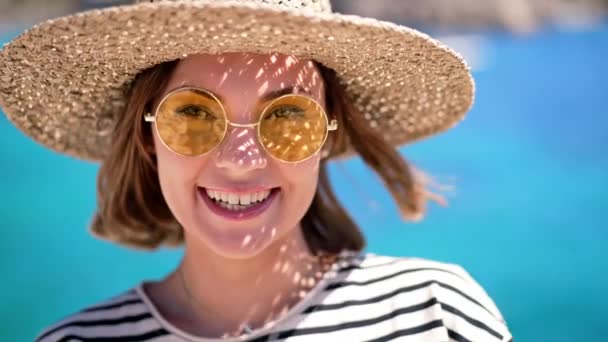 Sarı güneş gözlüklü ve hasır şapkalı güzel bir kadın gülümsüyor, deniz mavisi arka planında kameraya samimi bir şekilde gülüyor. Lüks seyahat yeri, tatil yaşam tarzı konsepti. — Stok video